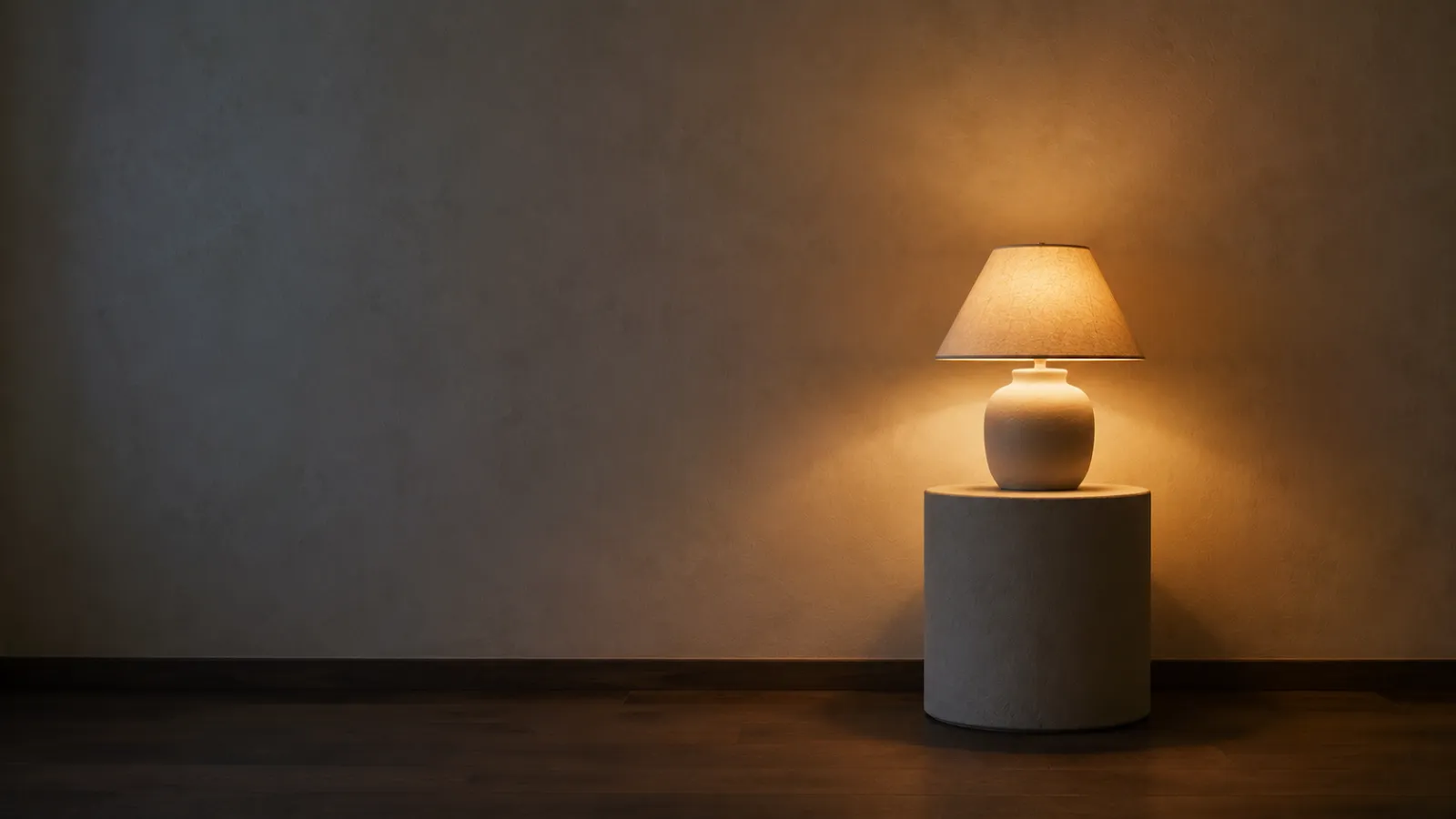 Lit ivory ceramic table lamp glowing on a plaster plinth in a dim room at dusk.