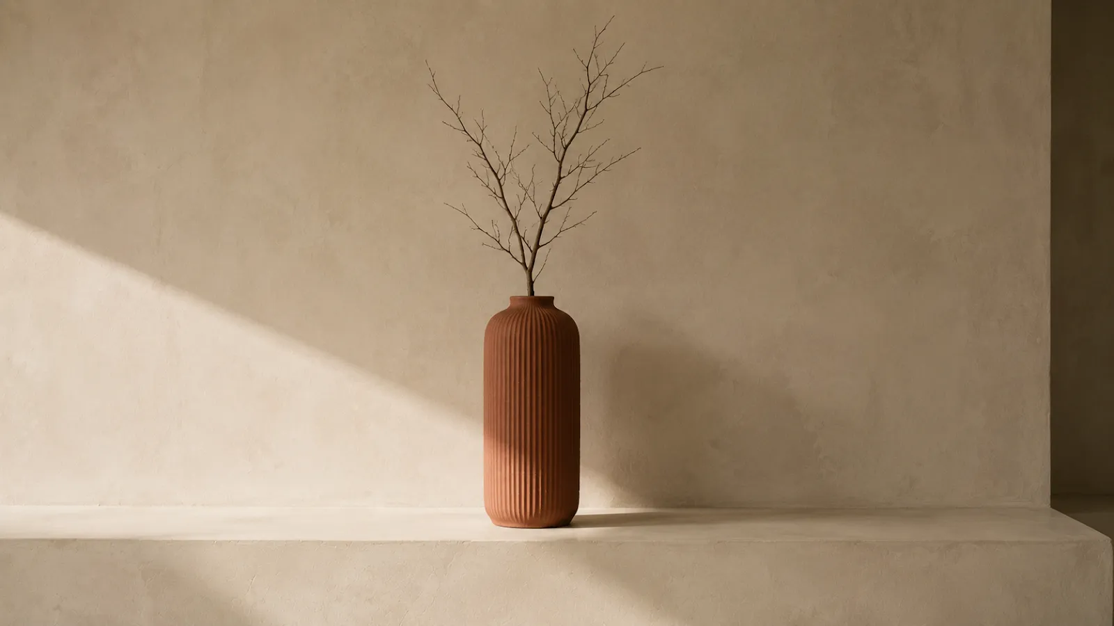 Ribbed terracotta vase with a single bare branch standing alone on a plaster ledge.
